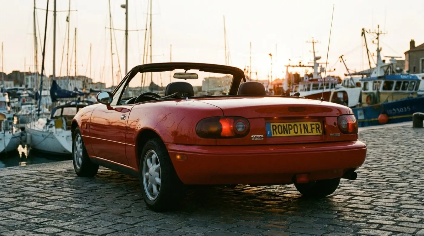 Mazda MX-5 NA rouge de trois quarts arrière capote ouverte près d'un port au coucher du soleil